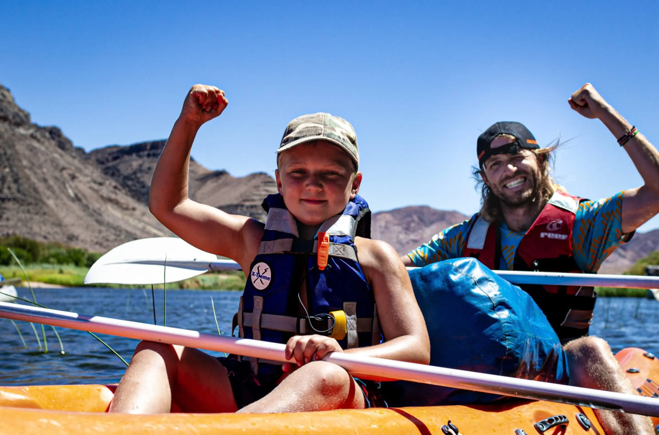 Een kind en een volwassene in een kajak op een rivier steken lachend hun armen omhoog in een overwinningspose. Ze dragen reddingsvesten en hoeden, met een blauwe lucht en rotsachtige heuvels op de achtergrond.
