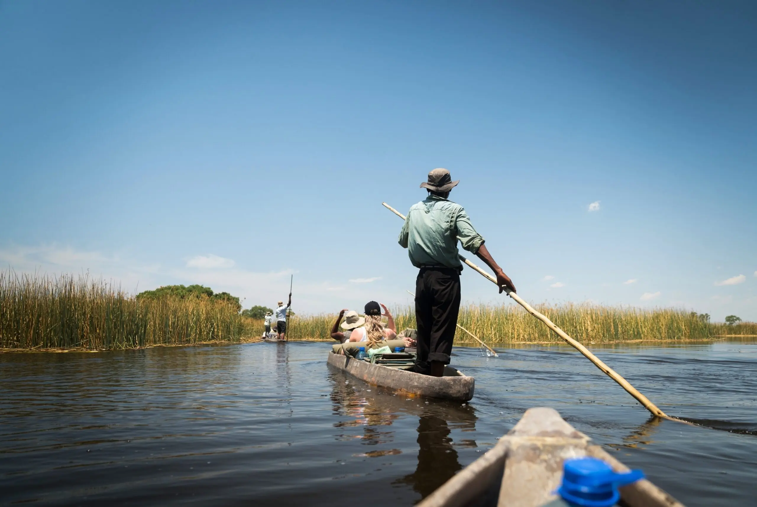 A person stands at the back of a wooden canoe, guiding it through calm water with a pole. Two passengers sit in front, surrounded by tall reeds under a clear blue sky. Another canoe is visible in the distance.