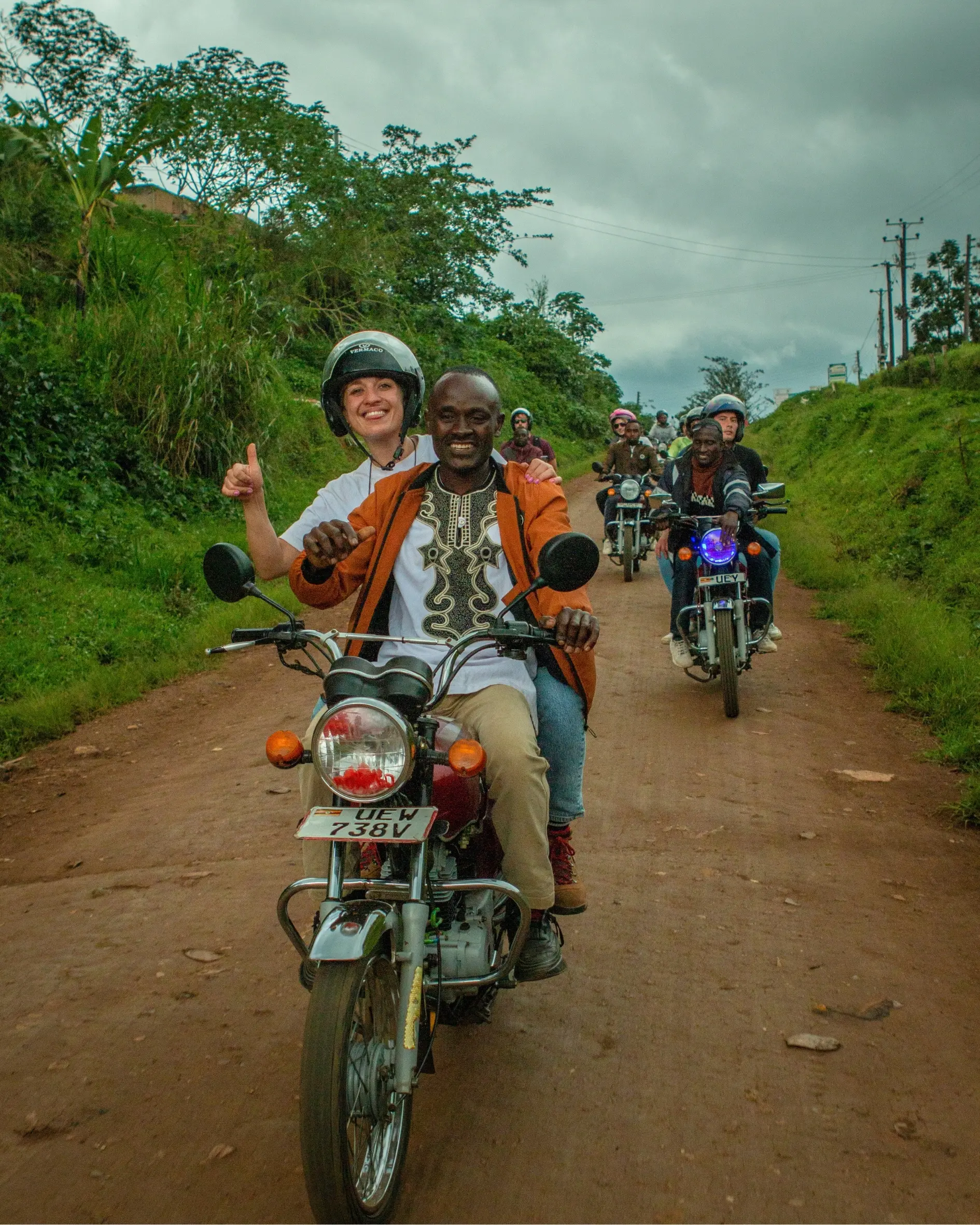 A group of smiling people ride motorcycles along a dirt road through green countryside; the front two riders, one wearing a helmet, give a thumbs-up gesture.
