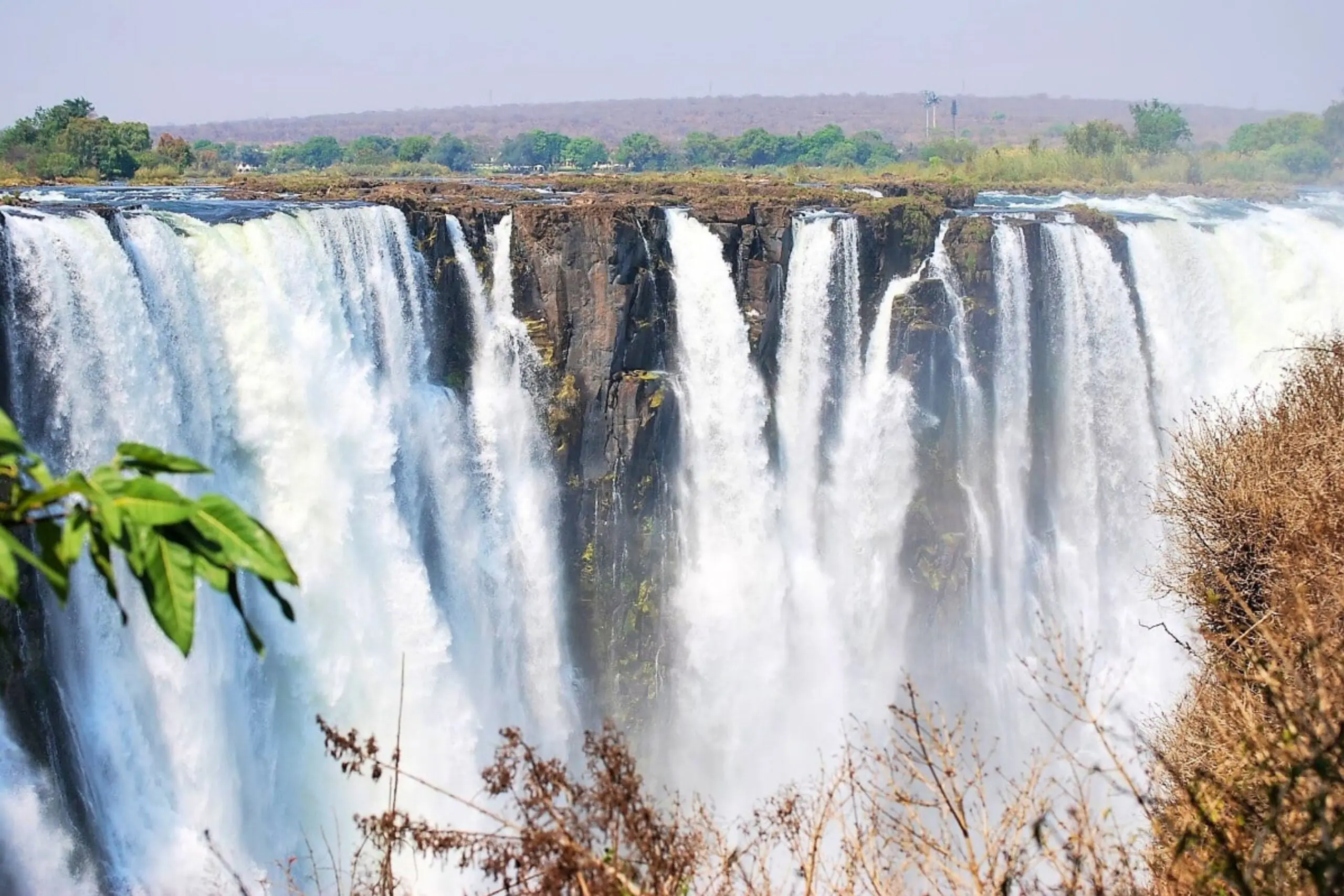 A wide view of powerful waterfalls cascading over a rocky cliff, surrounded by lush greenery and dry vegetation, with mist rising from below.