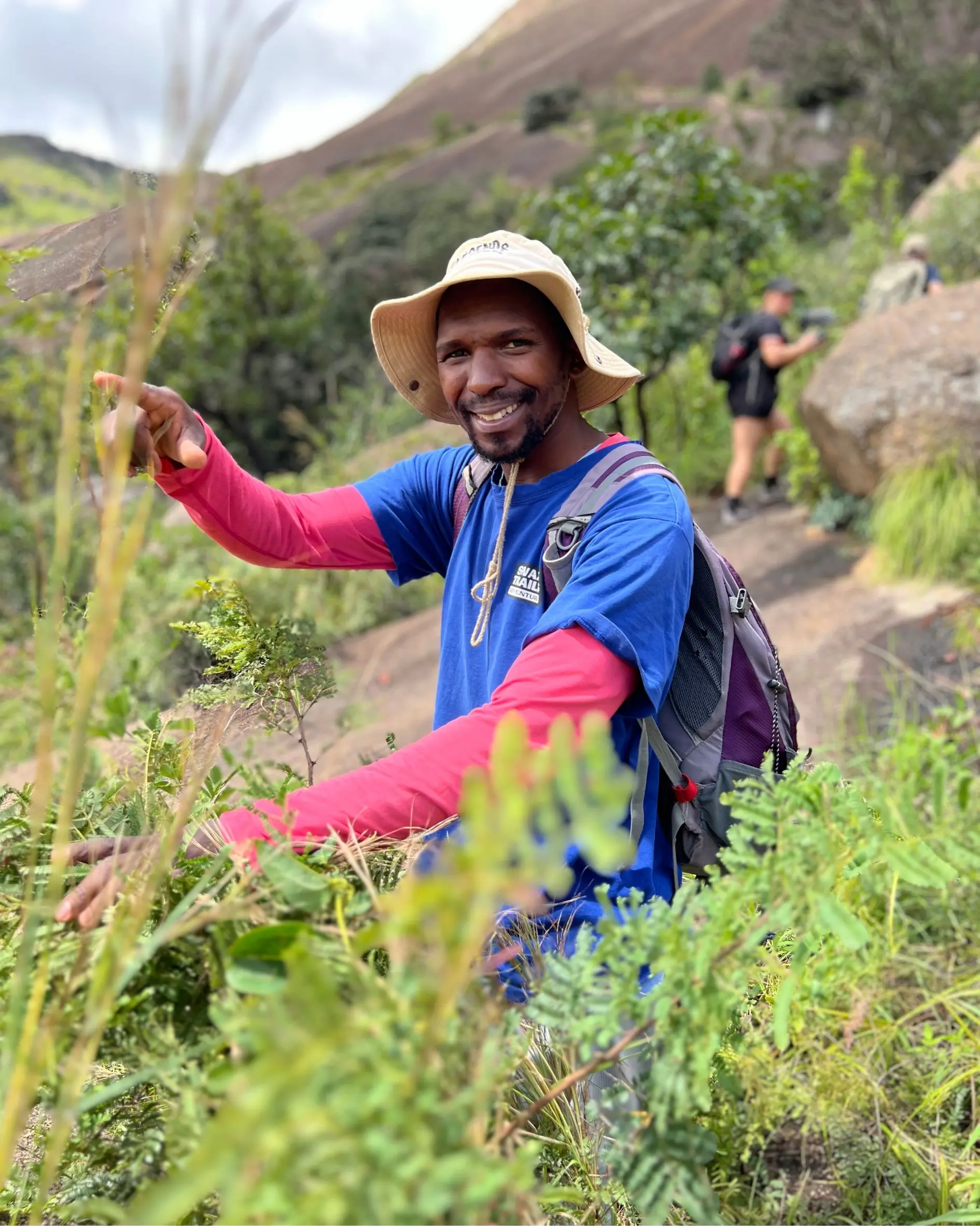 A smiling man in a blue shirt and wide-brim hat stands among green plants on a hiking trail, reaching out with one hand. There is another hiker in the background near rocks and trees.