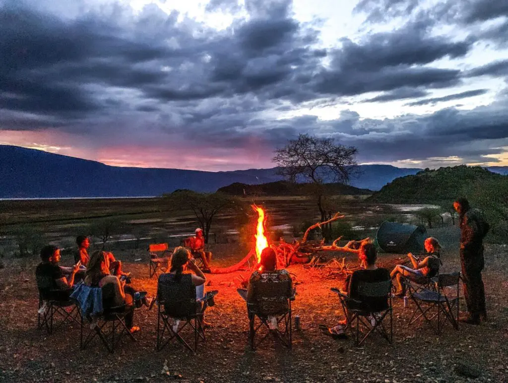 Een groep mensen zit in een cirkel van stoelen rond een kampvuur in de schemering, met dramatische wolken en een kleurrijke zonsondergang op de achtergrond, omringd door open wildernis en heuvels.