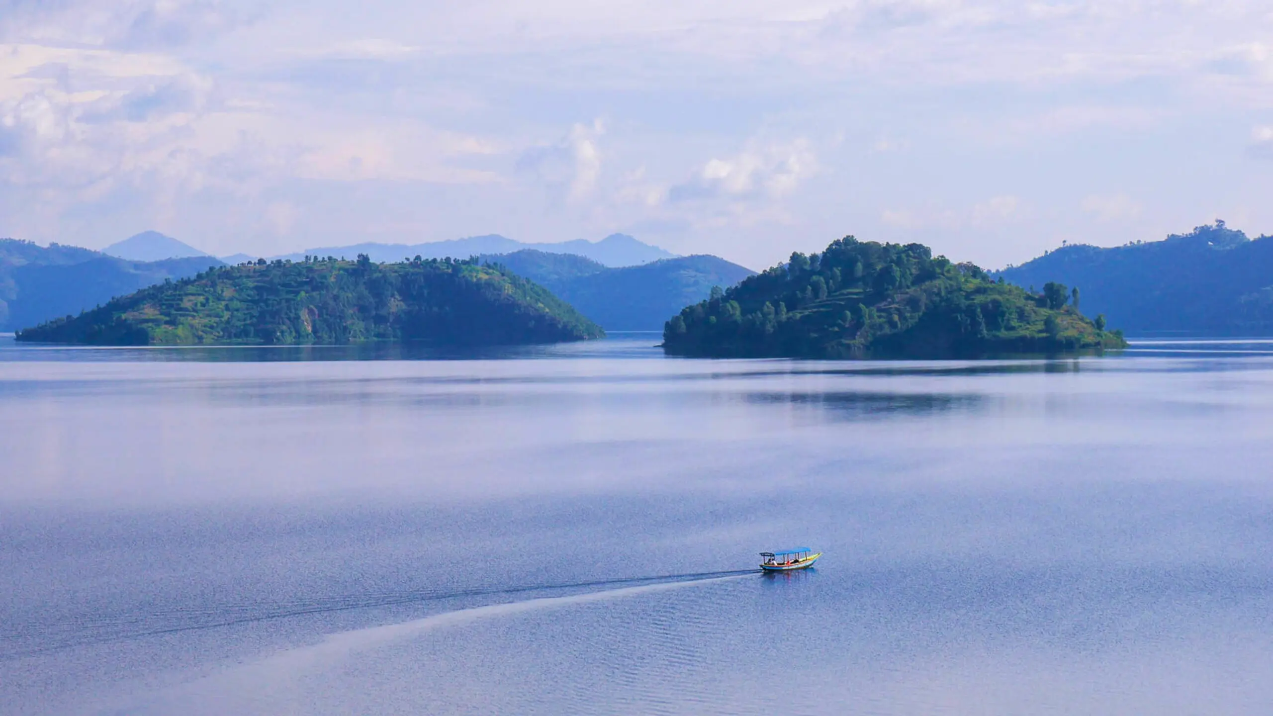 Een klein bootje vaart over een kalm blauw meer met twee groene, heuvelachtige eilanden op de achtergrond en bergen in de verte onder een gedeeltelijk bewolkte hemel.