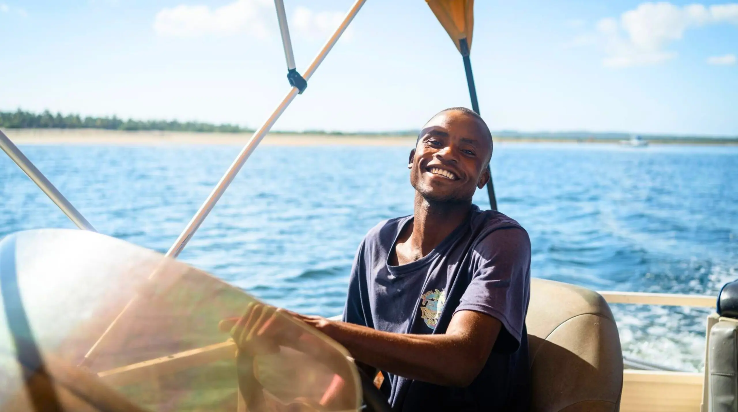 A smiling man steers a boat on a sunny day, with calm blue water and a distant shoreline in the background. He wears a dark T-shirt and sits at the helm under a canopy.