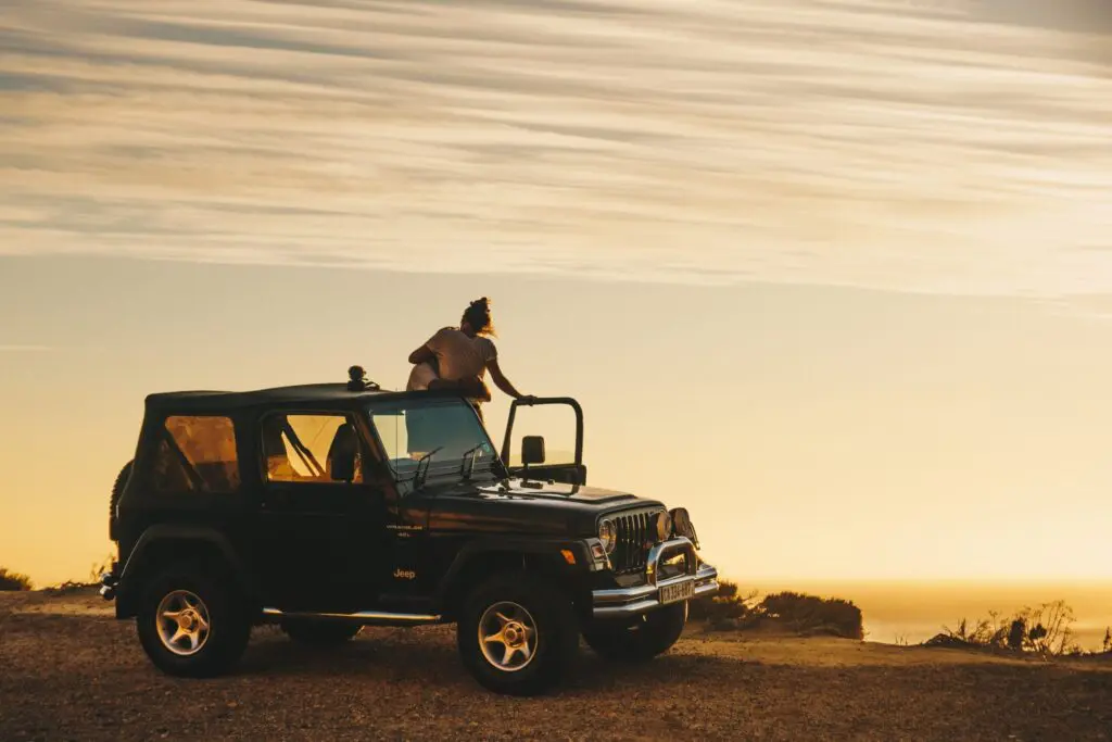 A person sits on the roof of a black Jeep parked on a dirt road in South Africa, gazing at a golden sunset with wispy clouds. The open landscape evokes a sense of adventure and freedom, perfect for anyone who loves travelling.