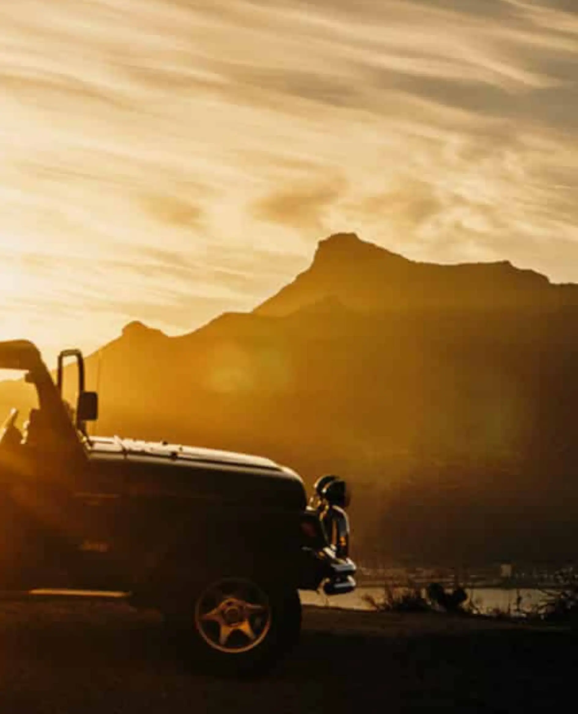 Een zwarte jeep geparkeerd op een weg bij zonsondergang, met een berg in silhouet op de achtergrond en een gouden hemel boven je hoofd.