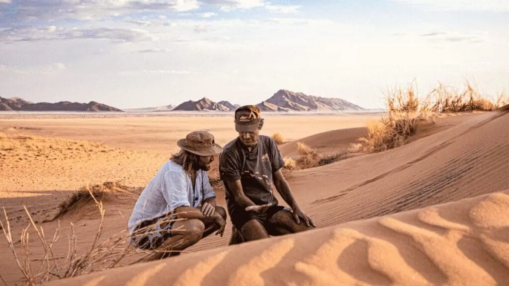 twee mannen bespreken iets op hun telefoon op een zandduin in namibië met uitgestrekt woestijnlandschap op de achtergrond