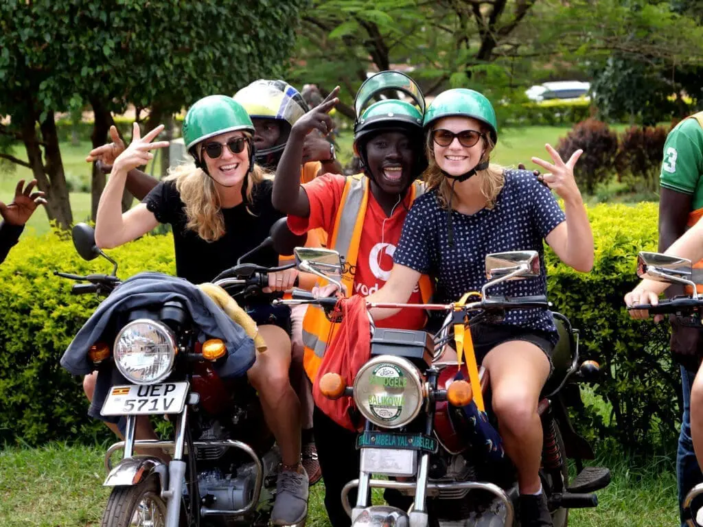 Drie lachende mensen met helmen op zitten buiten op een motor in Oeganda en poseren met vredestekens en handgebaren. Een persoon in een oranje vest staat tussen hen in, omringd door groene struiken en bomen - perfect voor Afrika reizen avonturen.