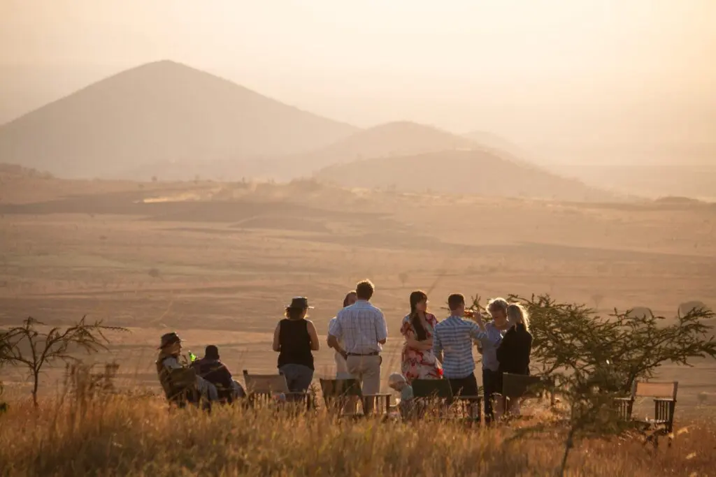 A group of people stand and sit together outdoors on a grassy plain at sunset in Tanzania, with rolling hills and mountains in the hazy background. The travelling scene is warm and relaxed, surrounded by sparse trees.