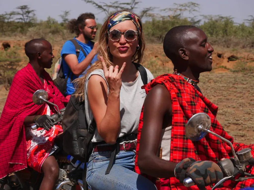 A woman in sunglasses and a headscarf makes a hand gesture while travelling on a motorcycle with a man in traditional Maasai attire. Another pair rides behind them, all enjoying the outdoors in rural Kenya, Africa.