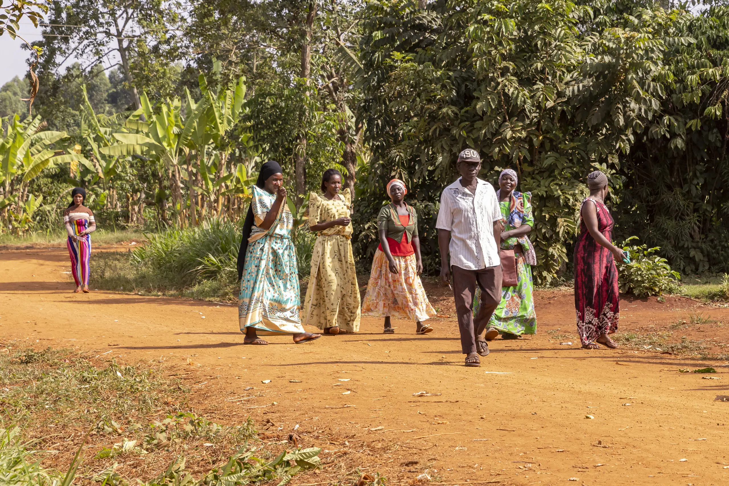 A group of people, mostly women in colorful dresses and one man in a white shirt, walk along a dirt path surrounded by green plants and trees in a rural setting. Another person stands further back on the path.