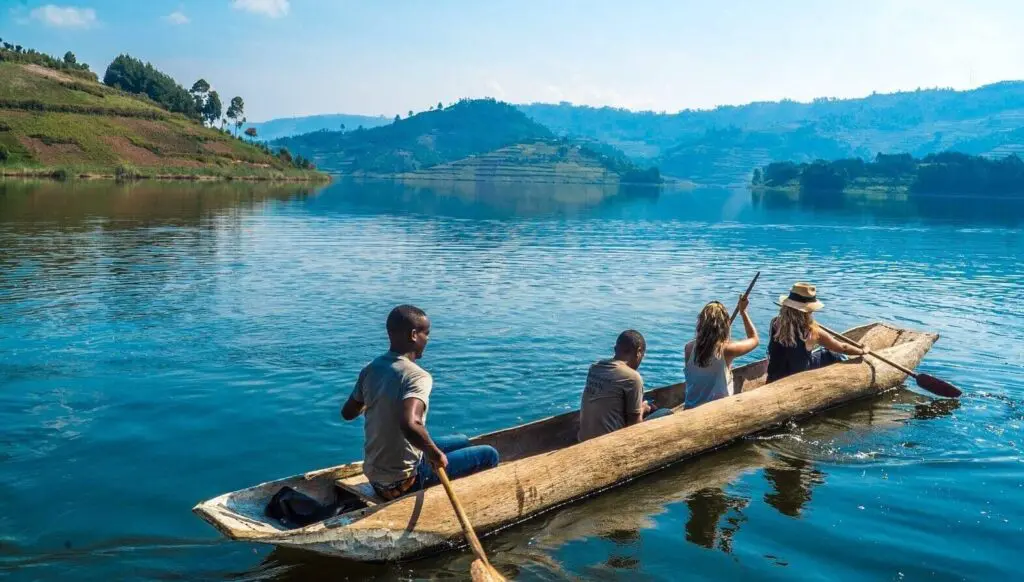 Four people paddle a wooden canoe across a calm lake in Uganda, surrounded by green hills and distant mountains under a blue sky. The scene is peaceful and sunny—a perfect travelling moment in Africa.