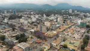 Luchtfoto van een stad in Tanzania met veel middelhoge gebouwen, straten met auto's en veel groen. Op de achtergrond zijn heuvels en een bewolkte hemel te zien - een perfect tafereel voor reizen-enthousiastelingen.
