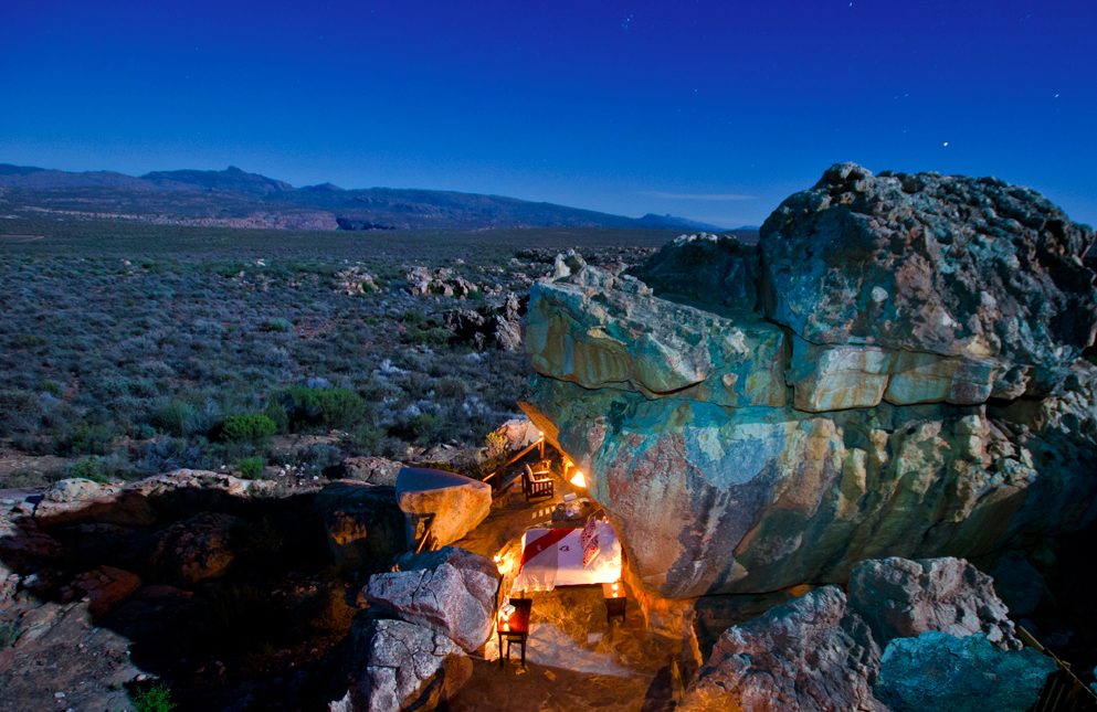 Een romantische eethoek buiten met een tafel voor twee is genesteld tussen grote rotsen, zacht verlicht door lantaarns, in een uitgestrekt, open landschap onder een heldere avondhemel.