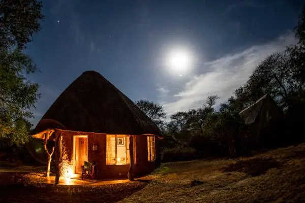 A round, thatched-roof hut glows warmly at night under a bright full moon and starry sky, surrounded by trees and natural landscape.