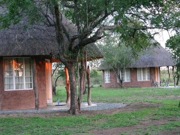 Two round huts with thatched roofs stand among green trees and grass. The huts have large windows, and soft light glows from inside one. The setting is peaceful and natural, suggesting a rural location.