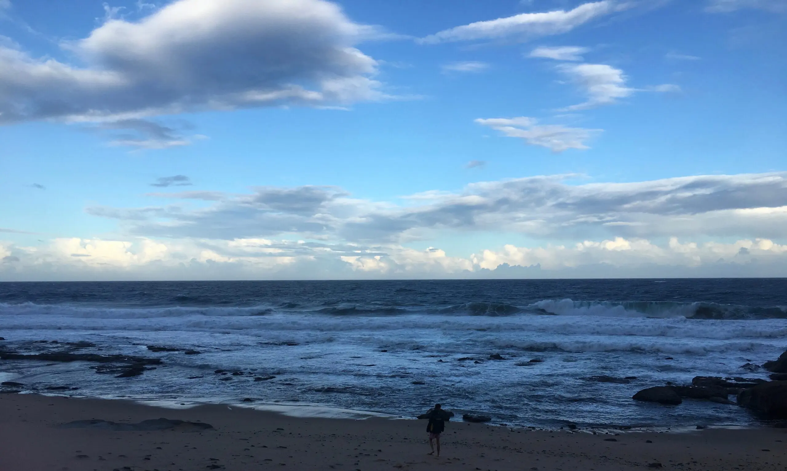 A person stands alone on a sandy beach near the shoreline, facing the ocean under a partly cloudy blue sky with waves crashing onto the shore.