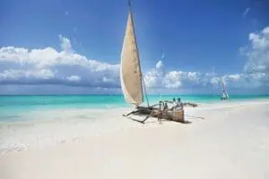 Een traditionele houten zeilboot met een beige zeil en meerdere mensen aan boord rust op de oever van een wit zandstrand in Tanzania, met turquoise water en een andere zeilboot in de verte onder een gedeeltelijk bewolkte hemel.