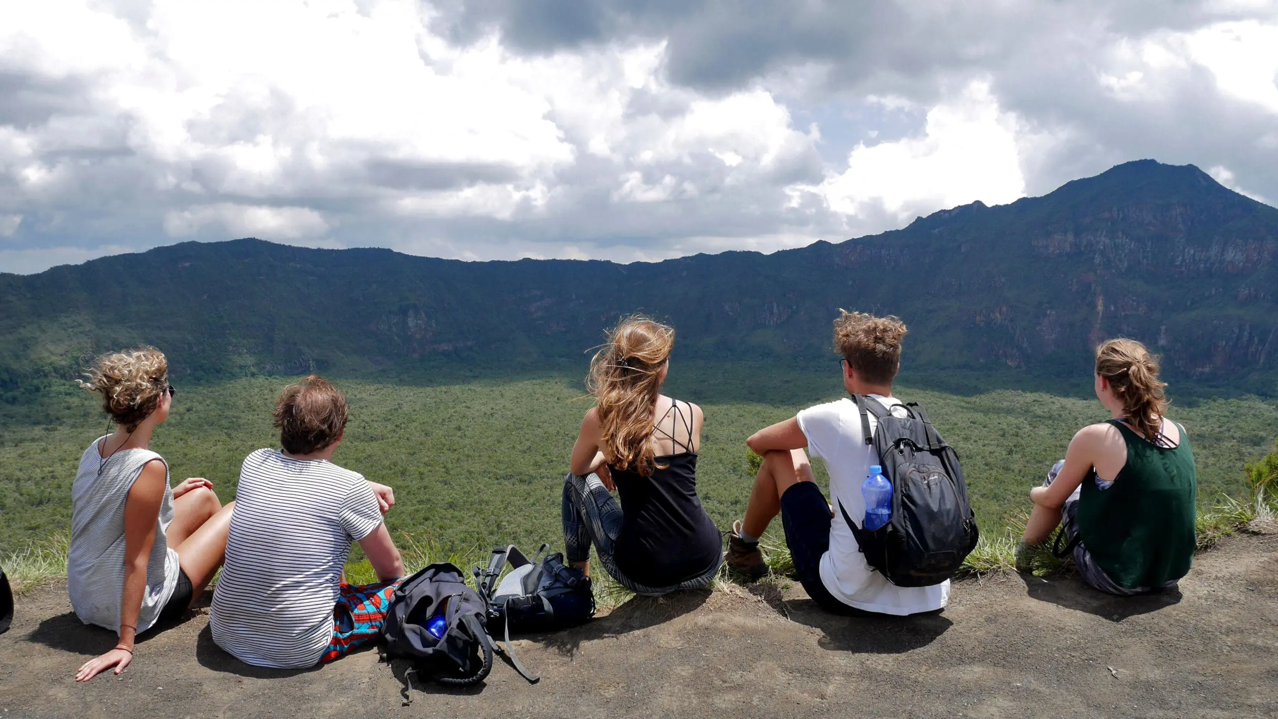 Five people sit on the edge of a cliff in Kenya, facing a vast green valley and distant mountains under a cloudy sky, with backpacks beside them—a perfect moment for travelling in Africa.