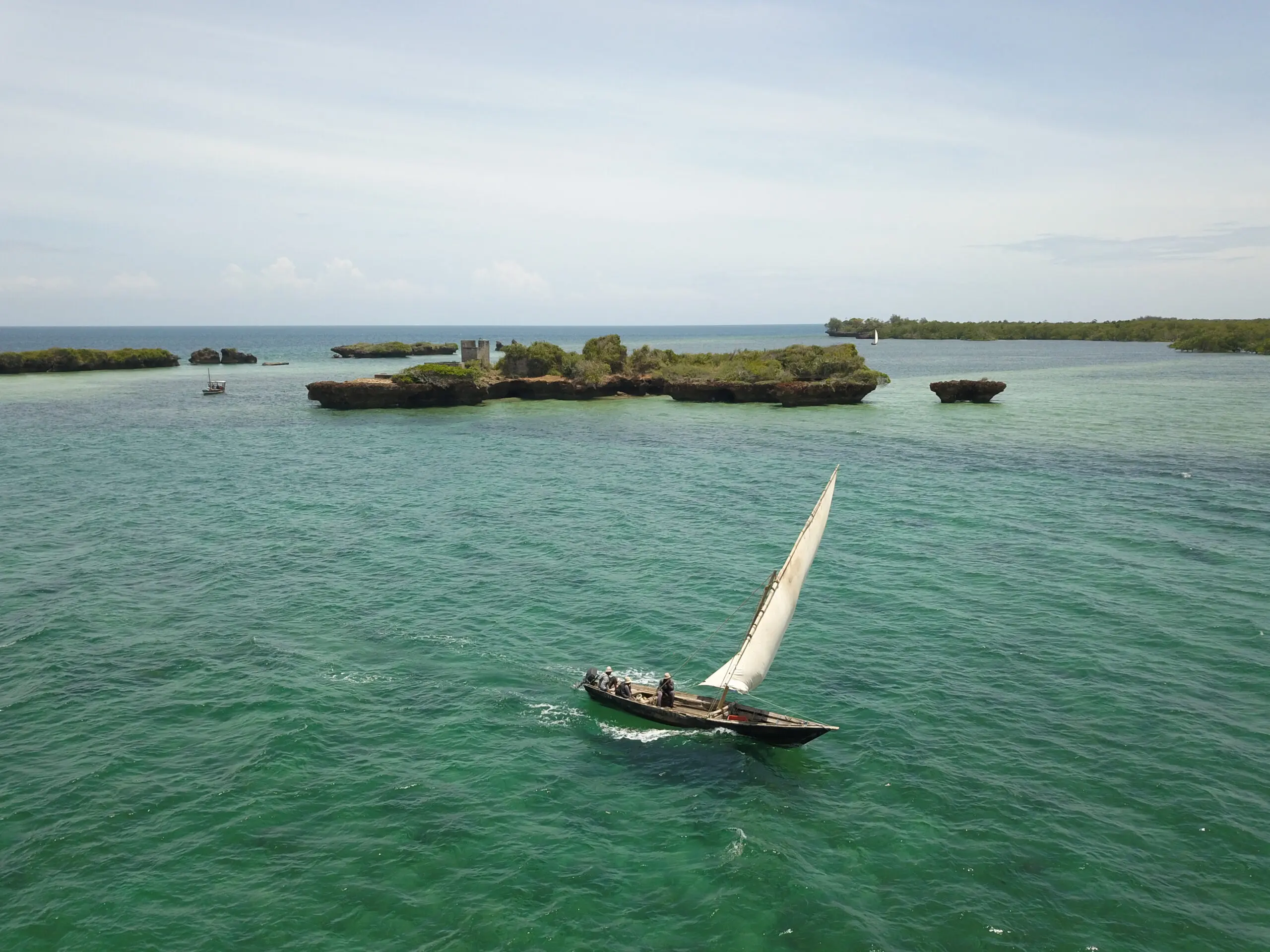 Een kleine zeilboot met mensen aan boord glijdt over turquoise water nabij rotsachtige, groene eilanden onder een heldere hemel.