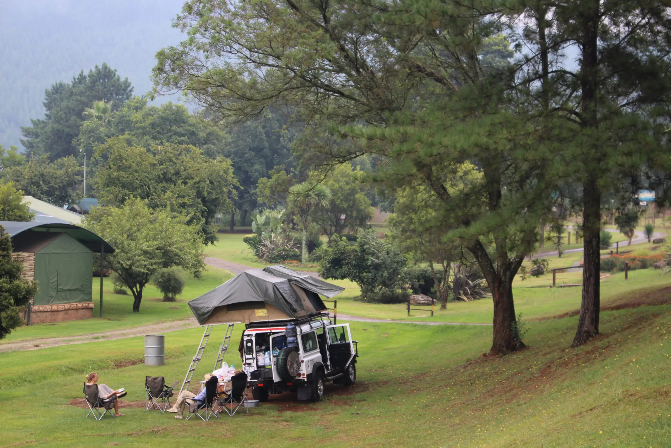 A group of people relax in camping chairs beside a white SUV with a rooftop tent in a grassy, wooded campsite surrounded by trees and greenery. Tents and forested hills are visible in the background.