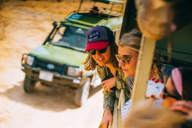 Twee mensen met zonnebrillen leunen uit een safarivoertuig, glimlachen en kijken voor zich uit. Op de achtergrond is een andere groene safari-jeep zichtbaar op een onverharde weg onder fel zonlicht.