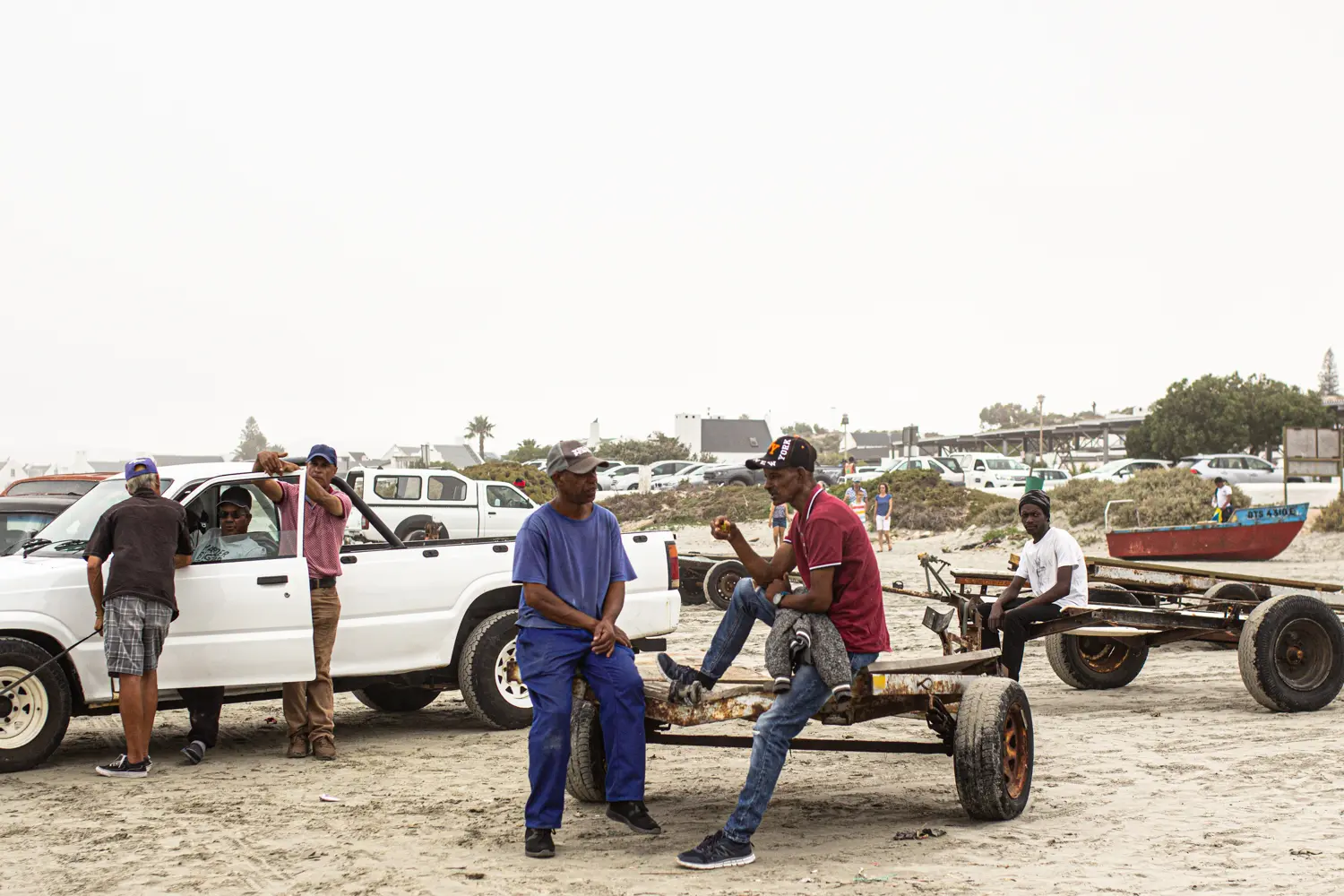 Several men gather on a sandy beach near white cars and makeshift carts. Two men sit and chat on a cart, while others stand or lean against a vehicle. A few buildings and trees are visible in the background.