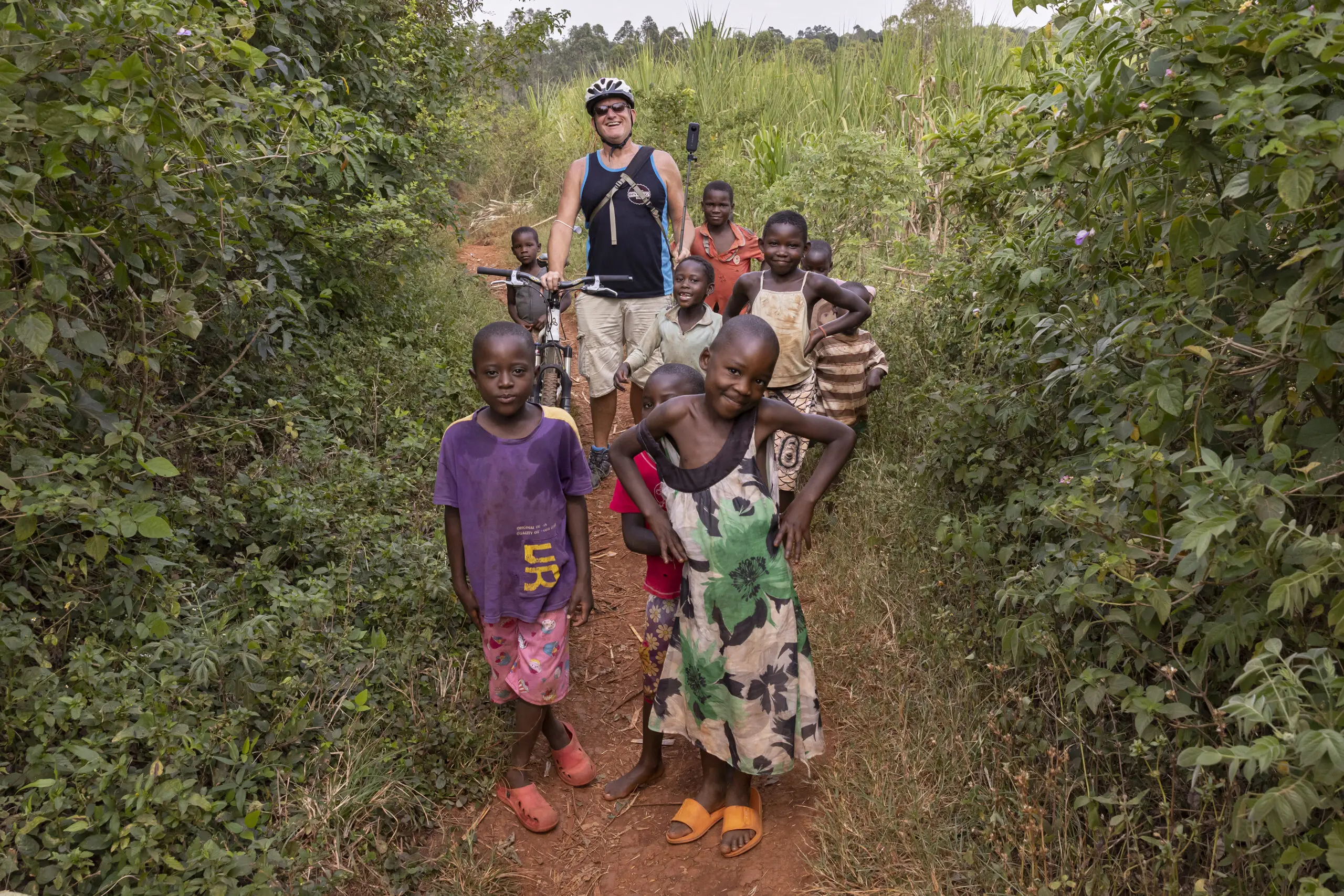 A group of smiling children stand on a dirt path surrounded by greenery, with a person wearing a helmet and sunglasses holding a bicycle and camera behind them. The children appear happy and playful.