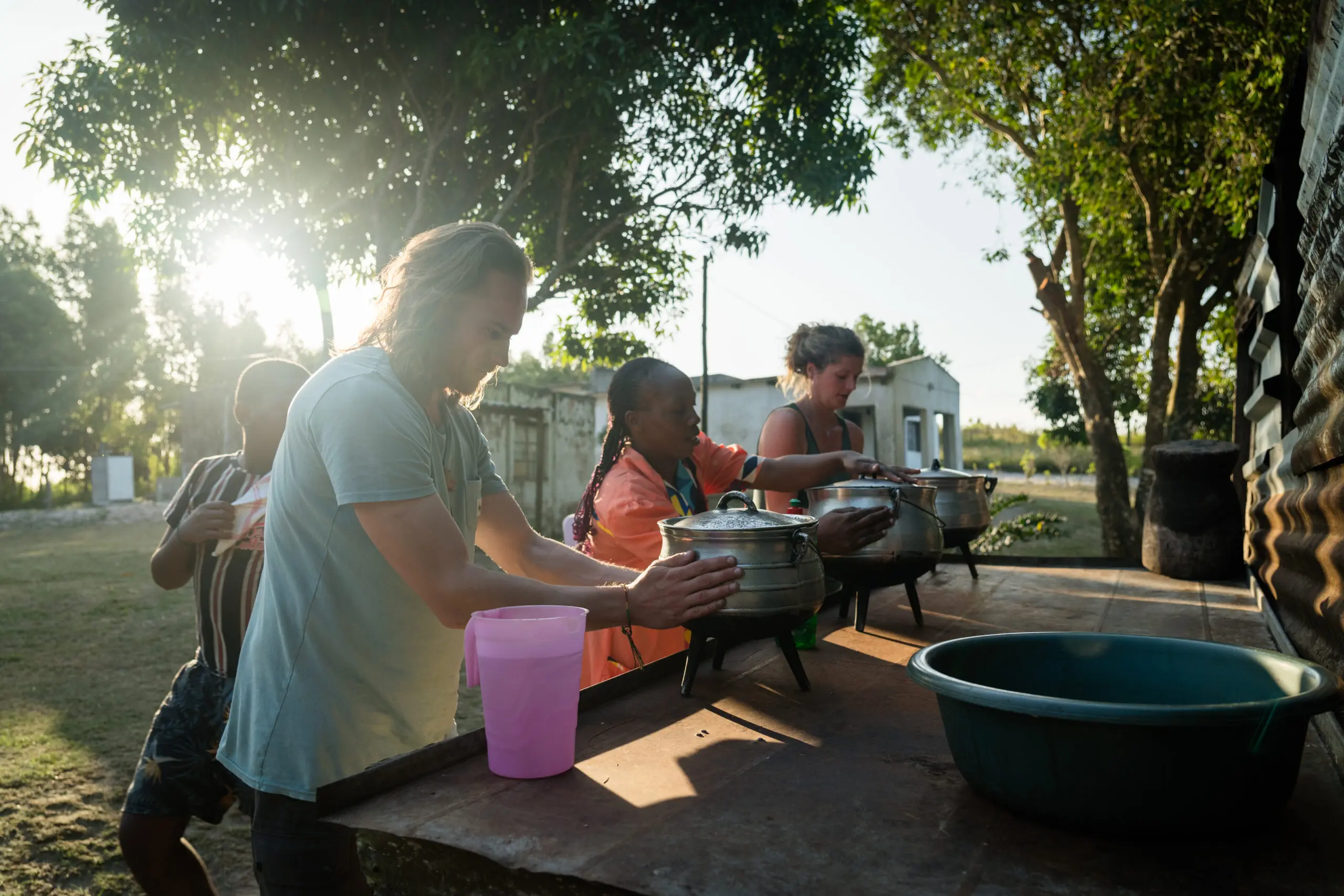 Three people stand outdoors at a long table, preparing food in pots under the sunlight. Trees and buildings are visible in the background, and a large green basin is on the table.