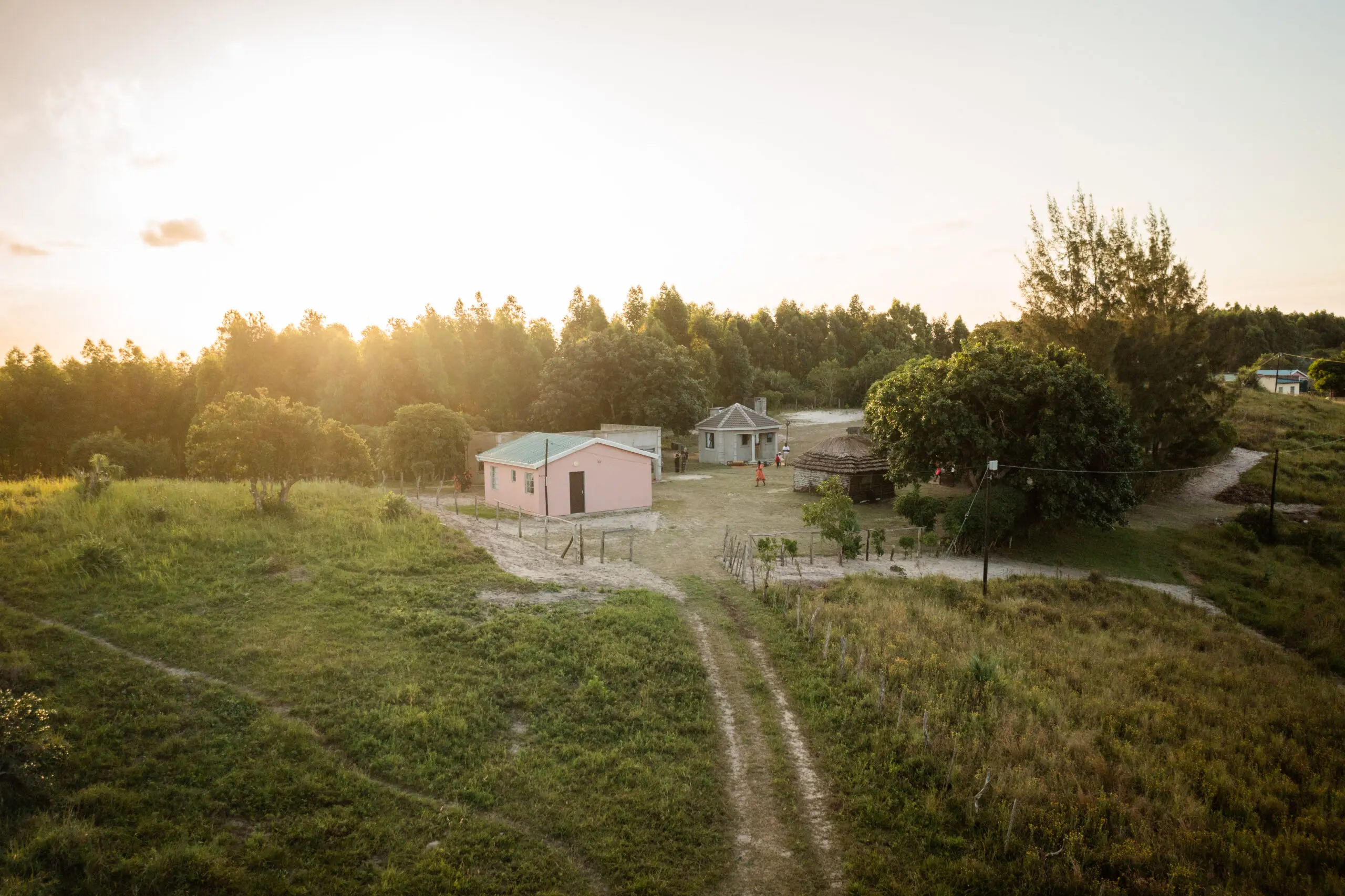 A rural landscape at sunset with a pink house, a smaller building, trees, and grassy fields. Dirt paths lead to the houses, and the sun sets behind a wooded area in the background.