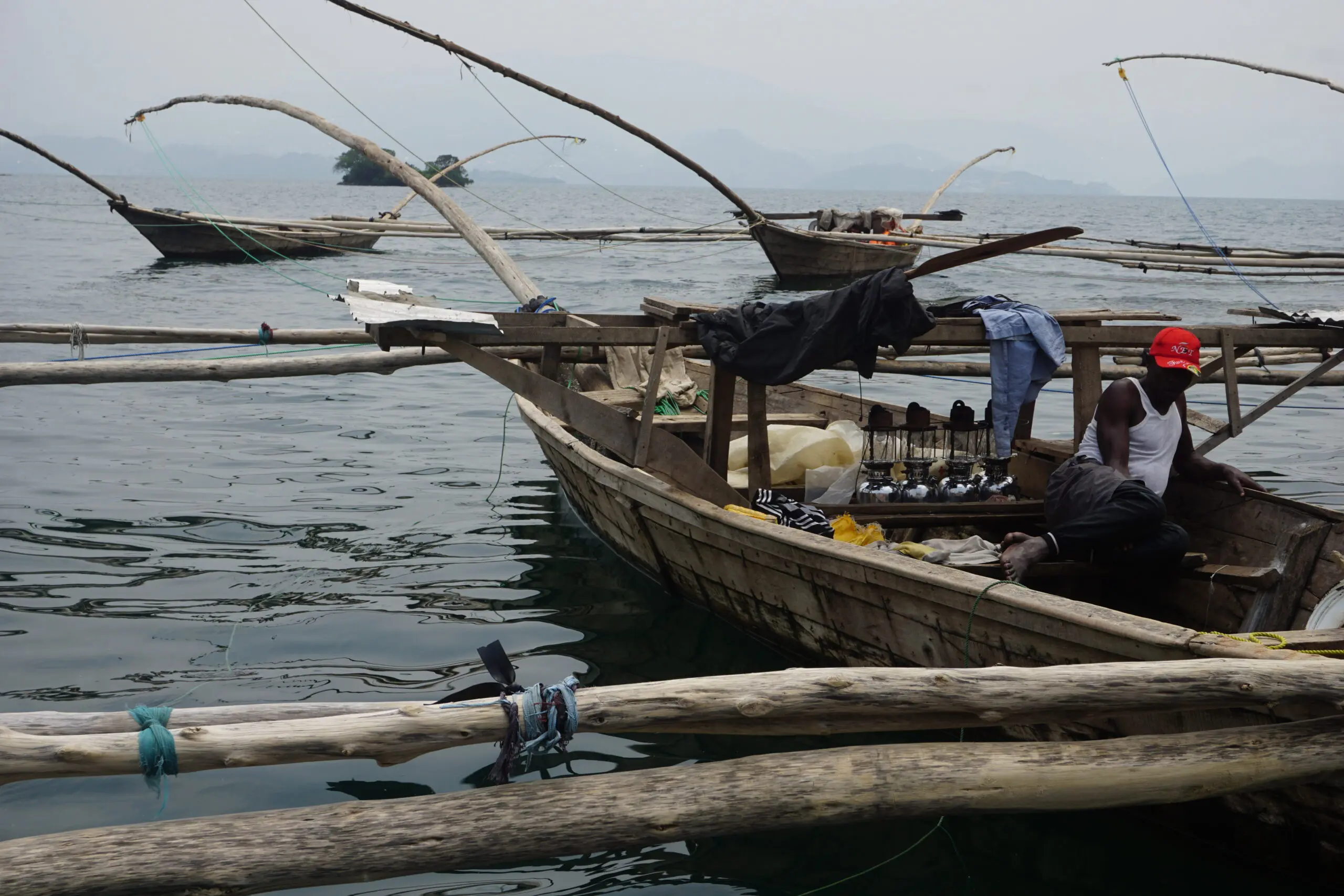 A man wearing a red cap sits on a wooden fishing boat surrounded by gear and clothing, with other similar boats floating on calm water and misty hills visible in the background.