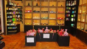 Een helder verlichte cadeauwinkel met planken met kleurrijke kaarsen en decoratieve voorwerpen. Tafels in het midden bevatten meer cadeaus, waaronder roze dierenfiguurtjes en dozen, allemaal netjes gerangschikt op donkere standaards.