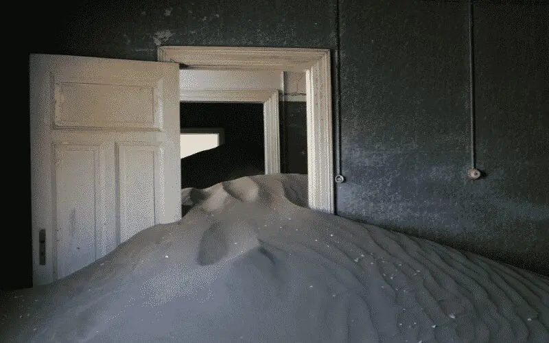 Ein mit Sand gefüllter Raum, der fast bis zur Oberkante einer weißen, offenen Tür reicht, während im Hintergrund noch mehr Sand durch die Türöffnungen rieselt. Die Wände sind dunkel und Drähte hängen herab und schaffen eine verlassene, surreale Atmosphäre.