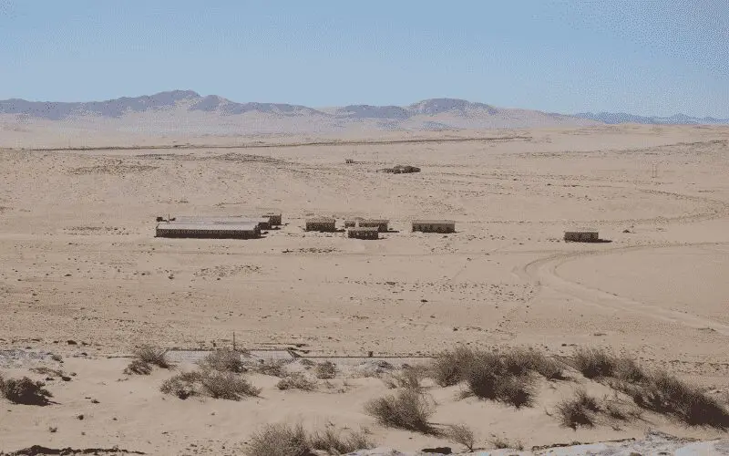 Een groep kleine, verweerde gebouwen staat geïsoleerd in een uitgestrekt woestijnlandschap onder een strakblauwe hemel, met bergen in de verte op de achtergrond en schaarse vegetatie op de voorgrond.