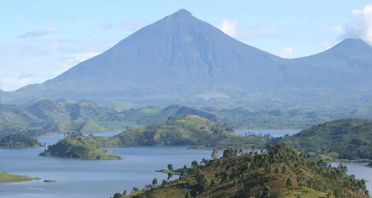 A scenic view of a large, green volcanic mountain towering over a lush landscape with rolling hills and a lake featuring several small forested islands under a partly cloudy sky.