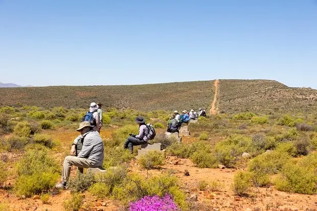 Mehrere Wanderer mit Hüten und Rucksäcken sitzen in Abständen auf Felsen oder Bänken entlang eines Weges in einer trockenen, mit Sträuchern bewachsenen Landschaft unter einem klaren blauen Himmel.