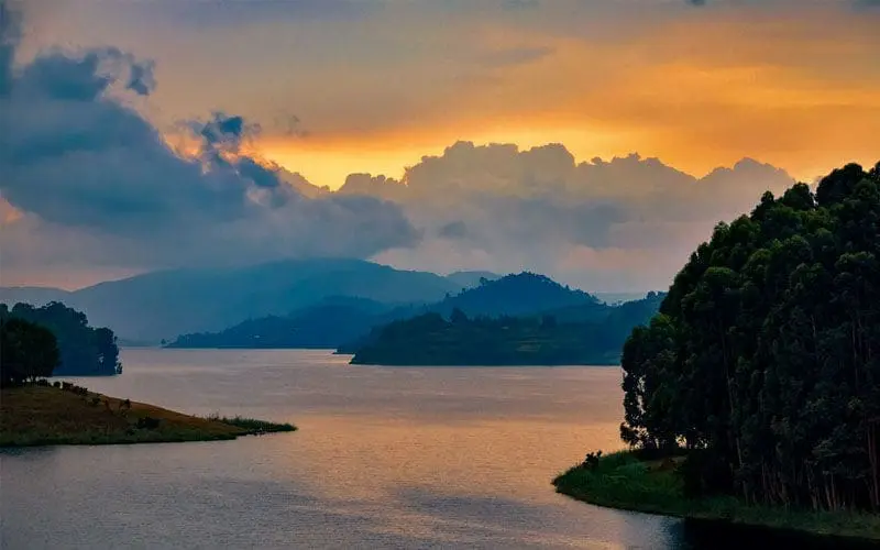 Een rustig meer bij zonsondergang in Afrika, met donkere silhouetten van bomen en heuvels. De dramatische lucht heeft gouden en blauwe tinten met dikke wolken - een uitnodigend tafereel voor wie van reizen houdt, geïnspireerd door de schoonheid van Oeganda.