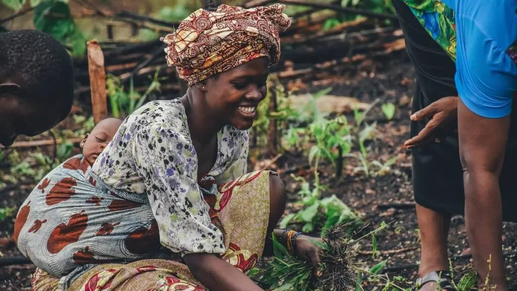 A woman wearing patterned clothing and a headscarf smiles while working in a garden in Kenya, holding plants. A baby is wrapped on her back, and other people are nearby, reaching toward the plants as they enjoy travelling through Africa.