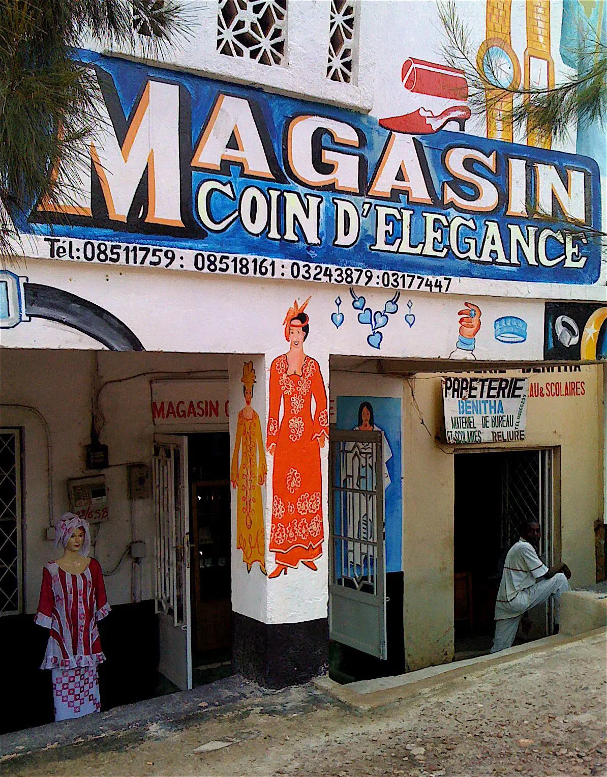 Een kleurrijke winkelpui in Rwanda met de naam MAGASIN COIN D'ÉLÉGANCE toont geschilderde afbeeldingen van elegant geklede vrouwen, met een persoon in een wit gewaad die een zijwinkel binnengaat. De bewegwijzering, rijk aan op Afrika geïnspireerde details, bevat telefoonnummers en decoratieve accenten.