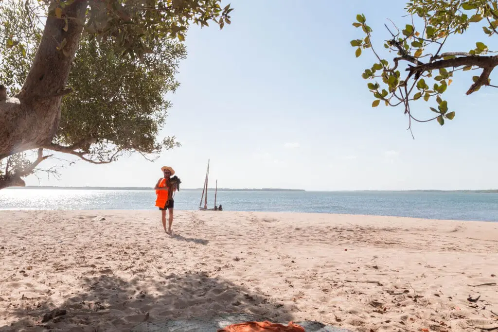reiziger in fel oranje reddingsvest loopt over wit zandstrand met dhowboot op de achtergrond aan de kust van lamu kenia