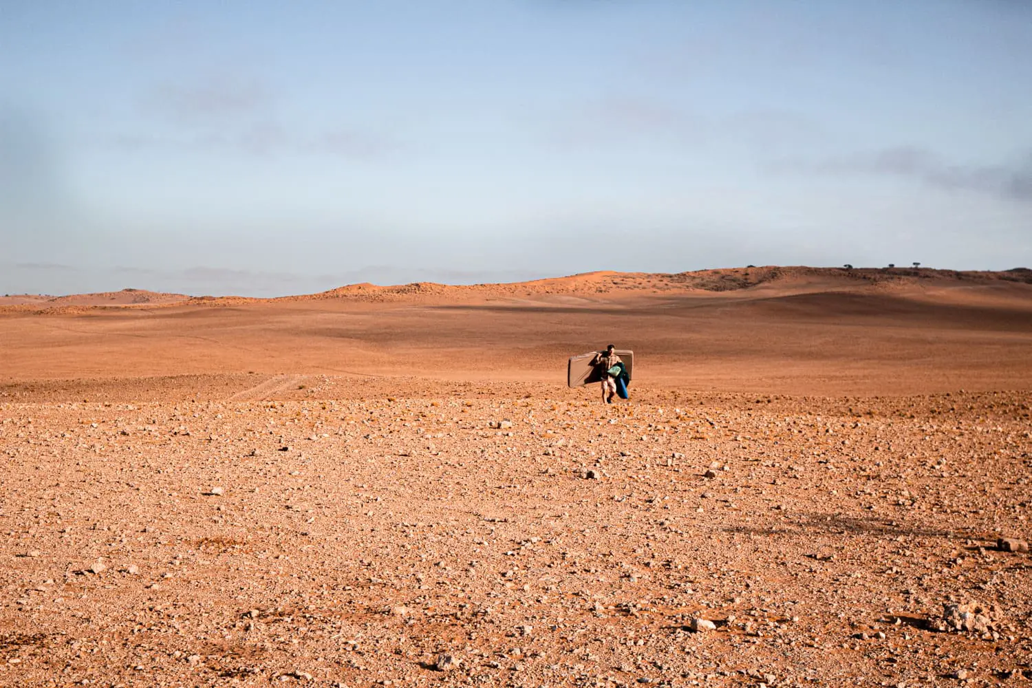een wandelaar loopt met kampeeruitrusting over de kale rotsbodem van een uitgestrekt woestijnlandschap in namibië