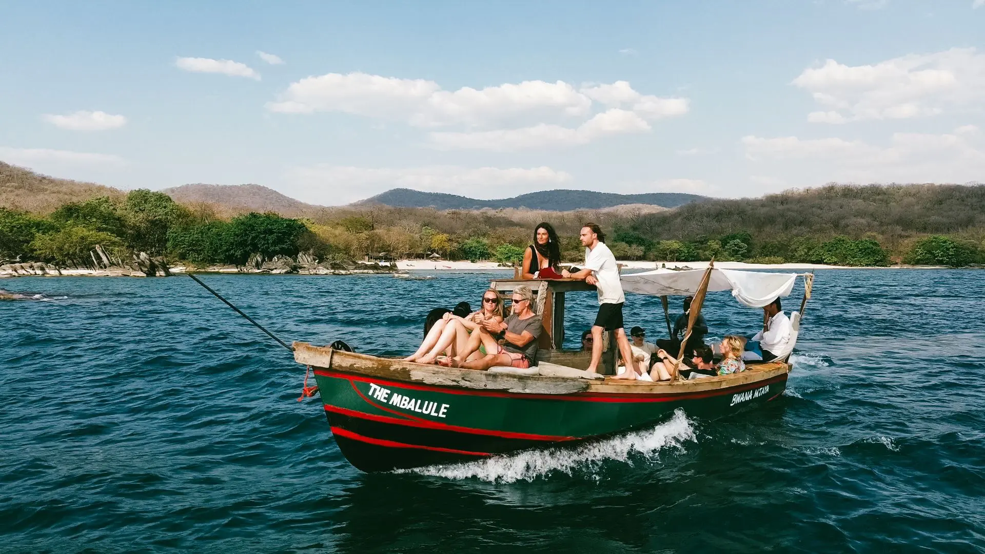 A group of people relax and smile on a small wooden boat named The Malulue, cruising on blue water near a tree-lined shore with hills in the background under a sunny sky.