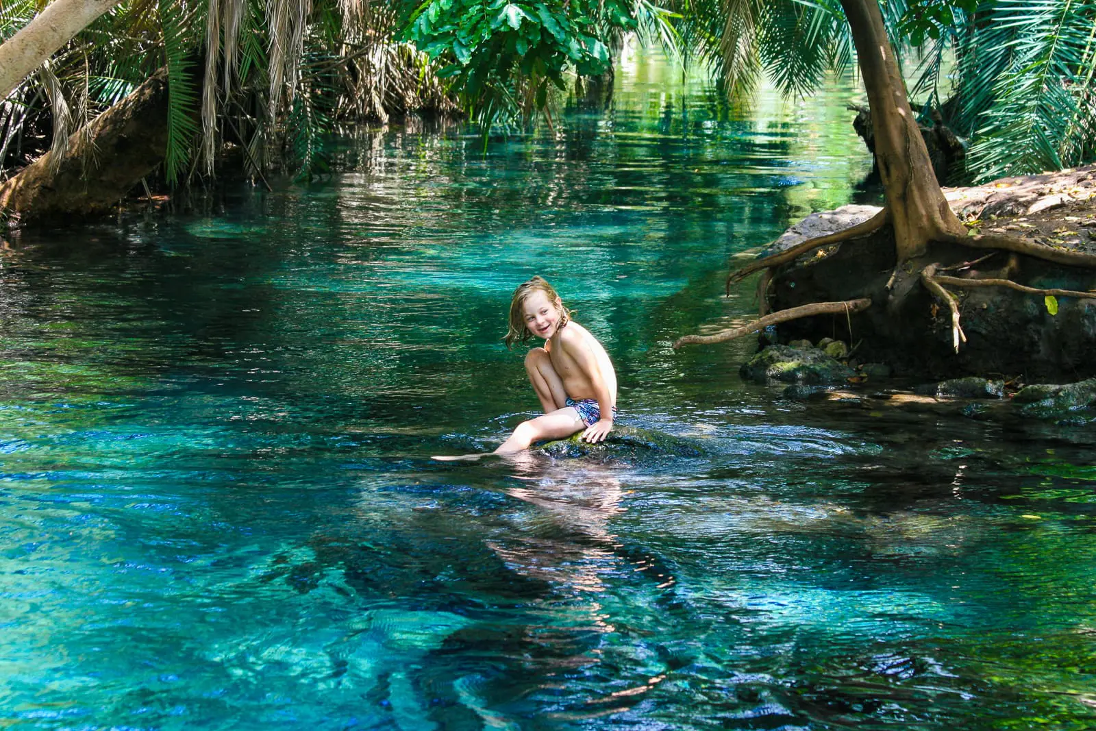 Een jong kind in zwemkleding zit en lacht op een rots in een helder, blauw natuurlijk zwembad omringd door weelderige groene bomen en planten - een beeld van de schoonheid van reizen door de prachtige landschappen van Afrika.