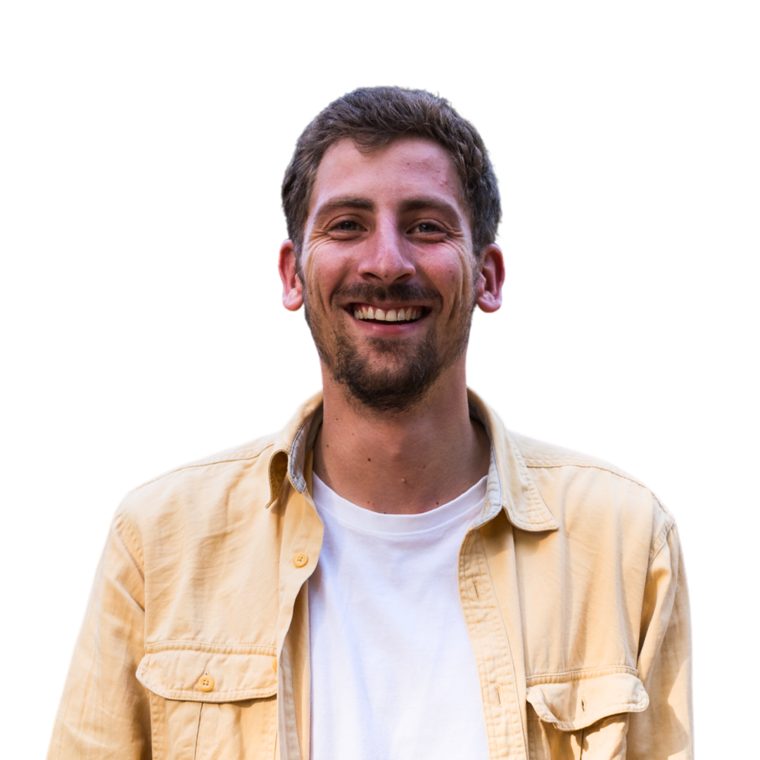 Een man met kort bruin haar en een baard glimlacht, draagt een lichtgeel overhemd over een wit t-shirt en staat tegen een effen witte achtergrond.