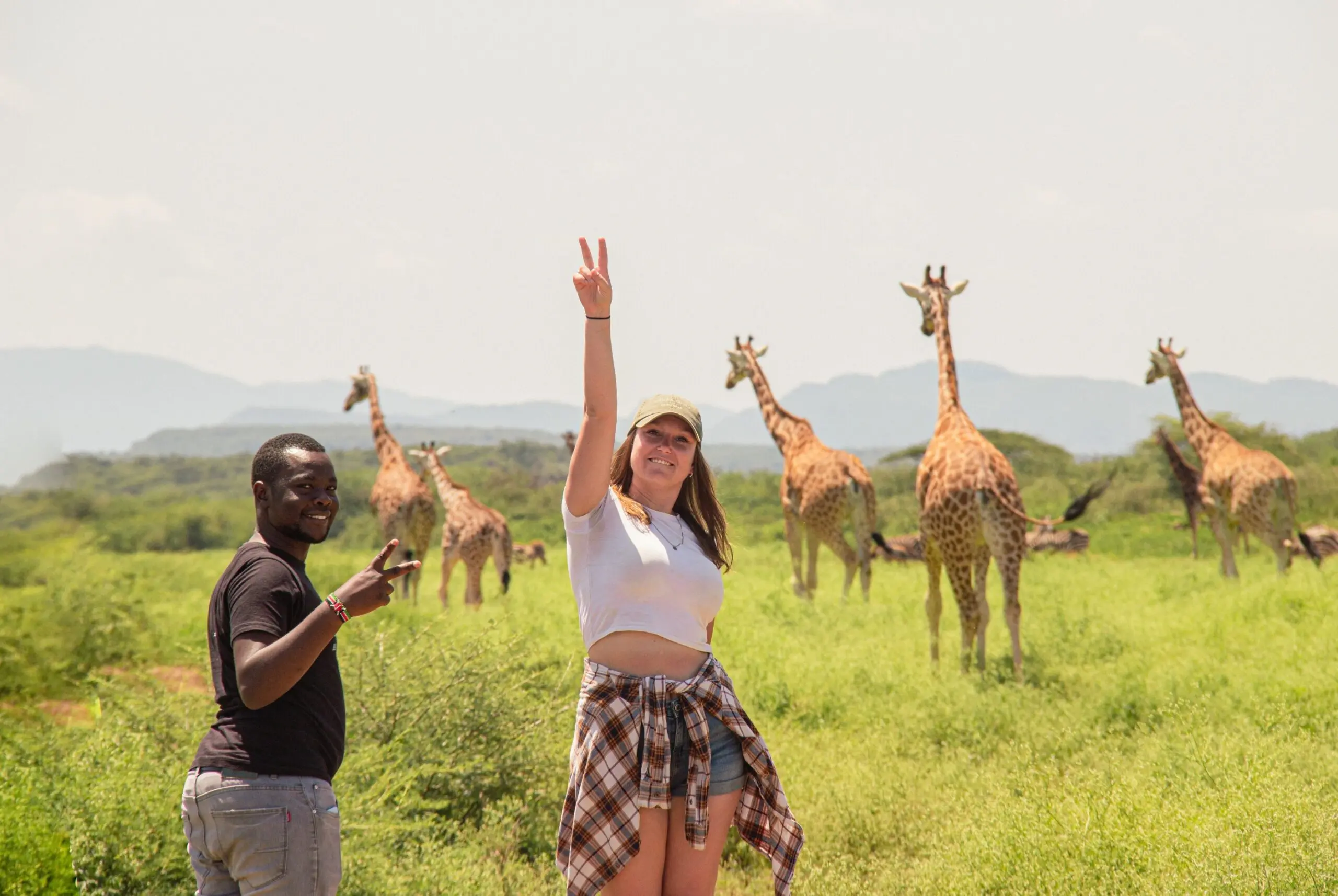 Twee mensen staan in een grasveld, glimlachen en maken vredestekens, met op de achtergrond een groep giraffen die grazen onder een zonnige hemel. In de verte zijn heuvels zichtbaar.