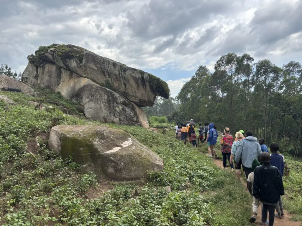 Een groep mensen loopt over een onverhard pad door groene vegetatie naar een grote, overhangende rotsformatie onder een bewolkte hemel, met hoge bomen op de achtergrond.