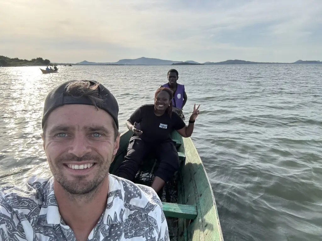 Drie mensen glimlachen terwijl ze in een kleine houten boot op een meer zitten met heuvels in de verte en een gedeeltelijk bewolkte lucht op de achtergrond. De voorste persoon neemt een selfie en één persoon maakt een vredesteken.