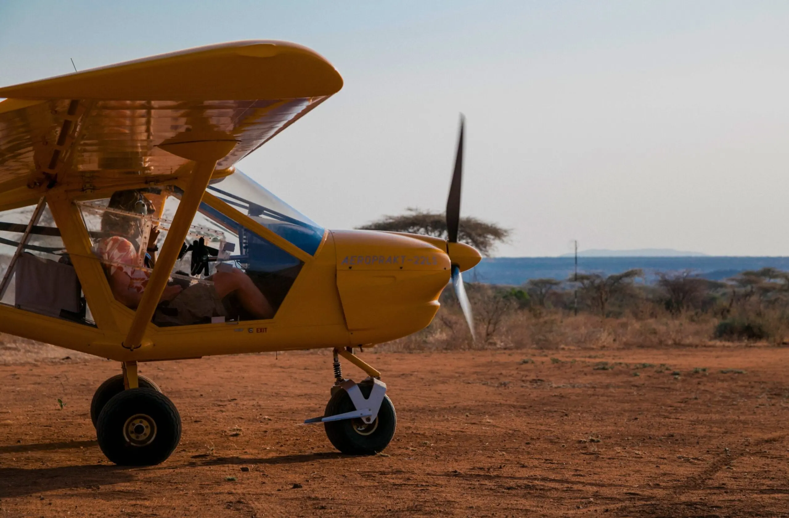 Een geel eenmotorig licht vliegtuigje met een persoon in de cockpit staat geparkeerd op een droge, roodachtige landingsbaan met schaarse vegetatie en bomen in de verte onder een heldere hemel.