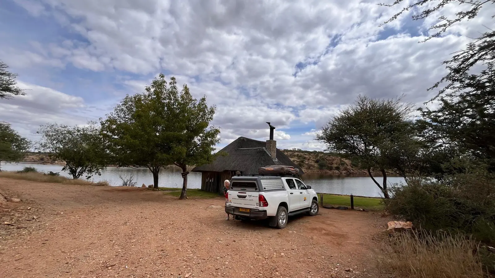 Een witte pick-up truck staat geparkeerd op een onverharde weg naast een rietgedekte hut bij een rustig meer, omringd door bomen en onder een bewolkte hemel.