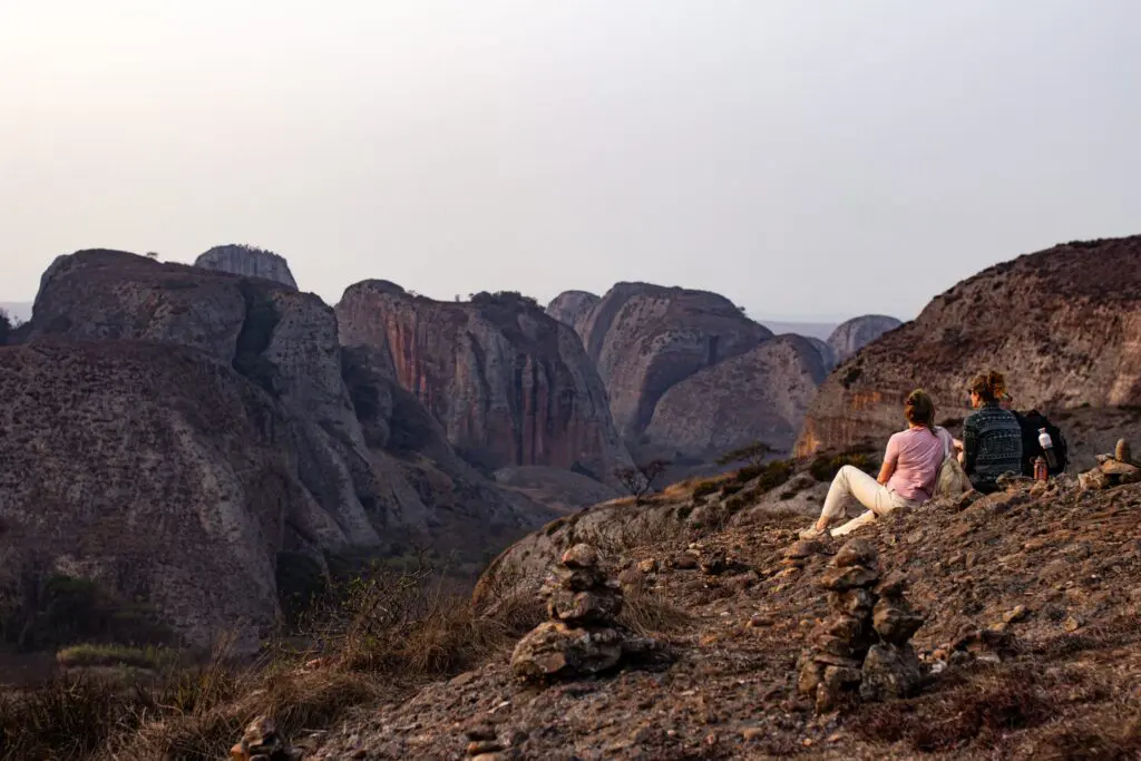 Twee mensen zitten op een rotsachtige heuvel en kijken uit over grote, ruige rotsformaties en kliffen onder een wazige hemel, met kleine stenen stapels op de voorgrond.